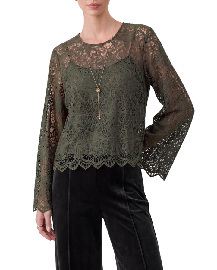 A woman modeling a dark olive green sheer lace long-sleeve top with a scalloped hem, paired with black velvet pants, against a solid gray studio background.