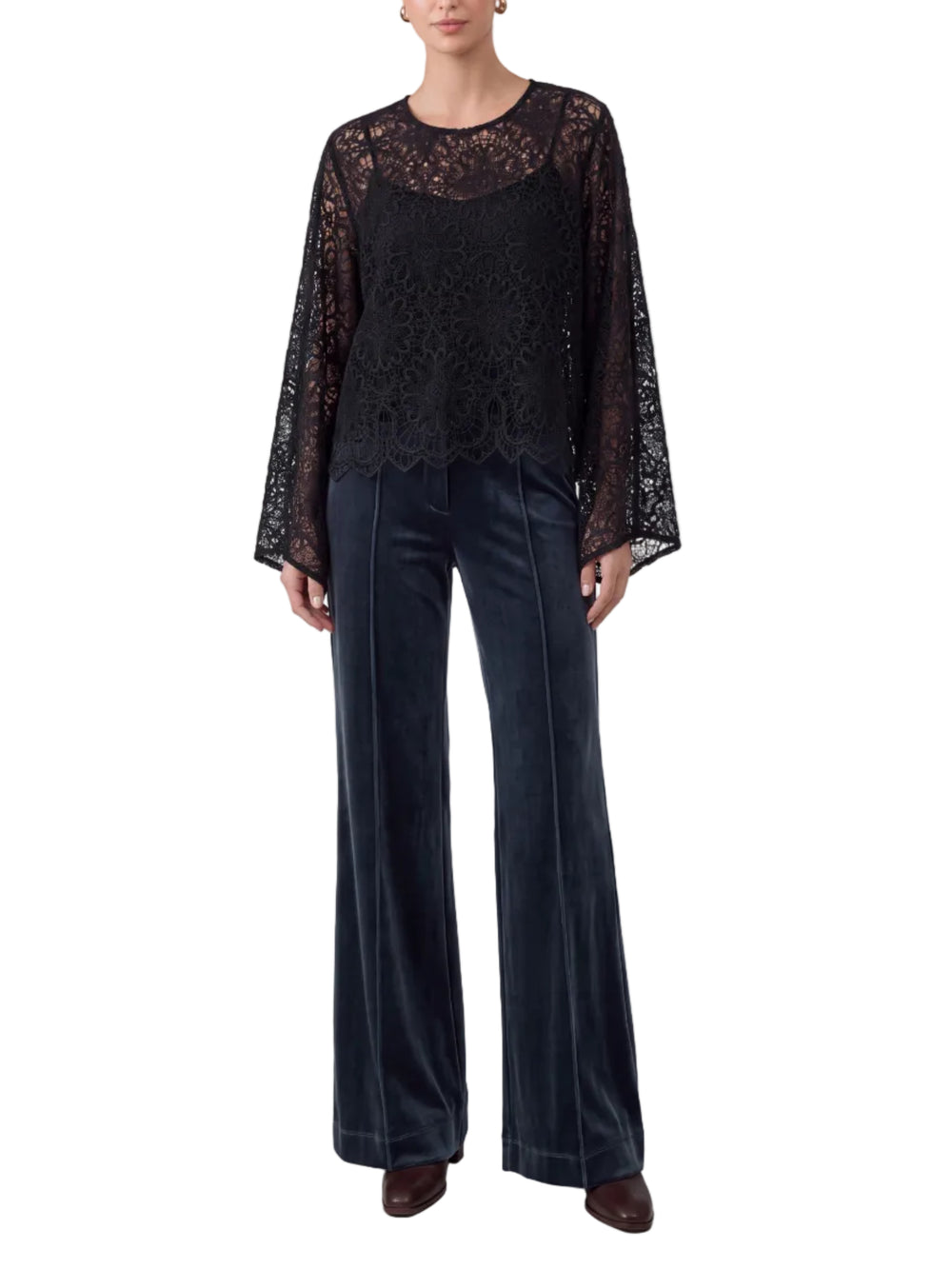 A woman stands against a light gray studio background wearing a black lace long-sleeve top with sheer flutter sleeves and dark navy wide-leg velvet pants.
