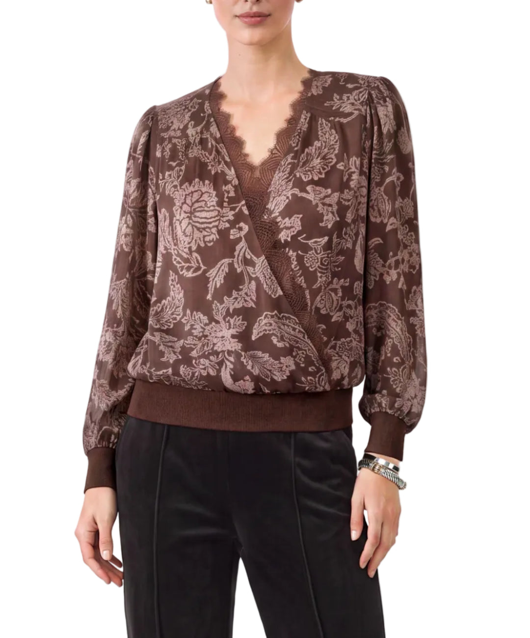 A woman modeling a brown long-sleeve blouse with a floral jacquard pattern, a scalloped V-neckline, and puffed shoulders, paired with black trousers against a light gray studio background.