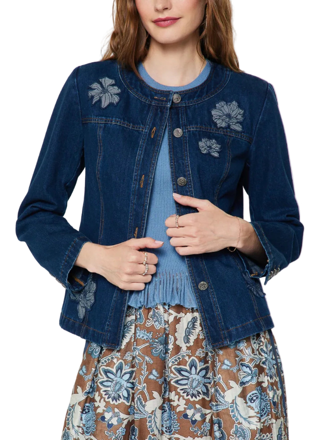 A woman wearing a dark blue denim jacket with floral embroidery on the chest, layered over a light blue top and a brown floral skirt, against a plain light gray background.