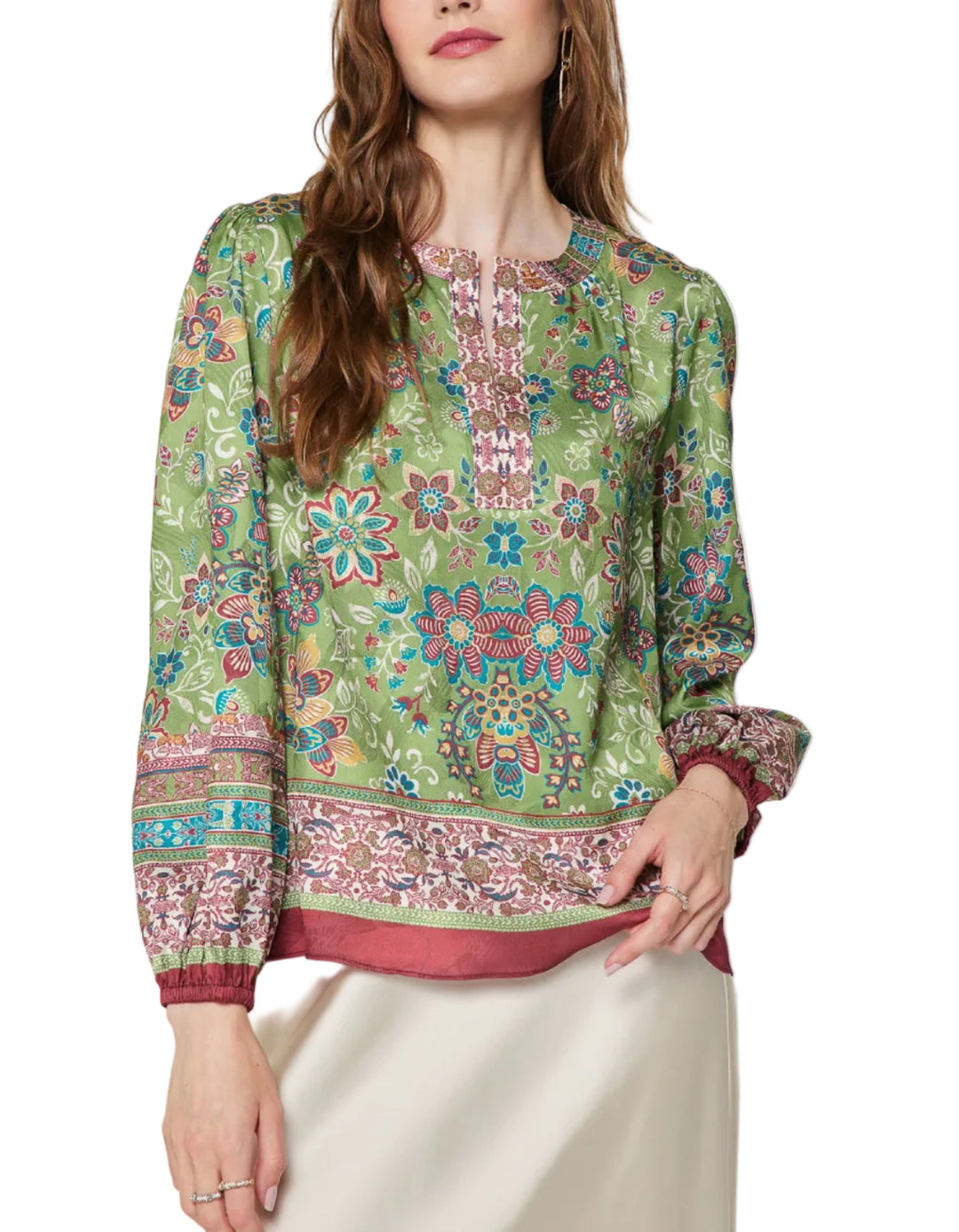 A woman wearing a green long-sleeve blouse with a colorful floral and paisley print, paired with a cream-colored skirt, against a light gray studio background.
