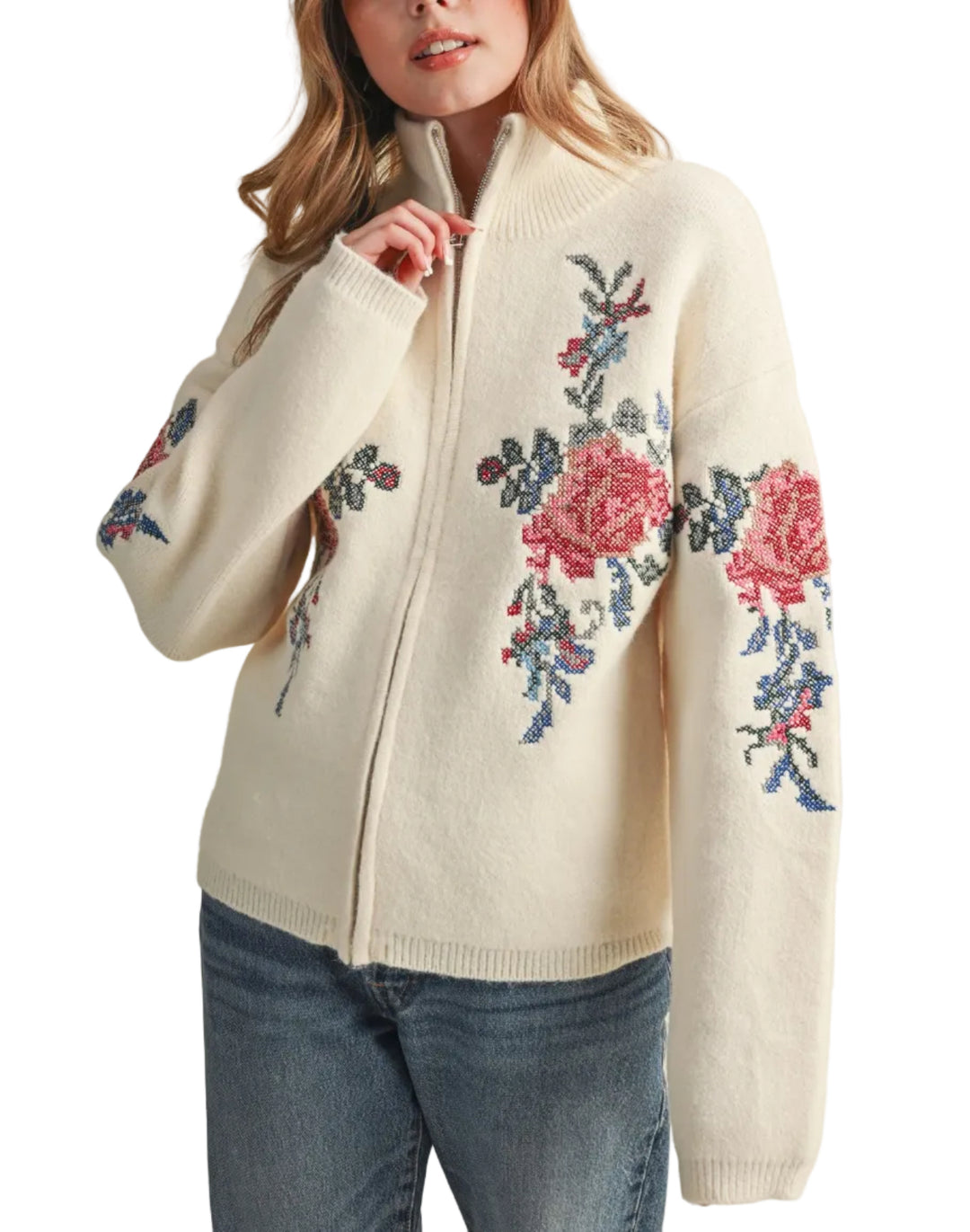 A woman wearing a cream-colored knit zip-up sweater with colorful floral embroidery on the front and sleeves, paired with blue jeans, set against a plain beige background.