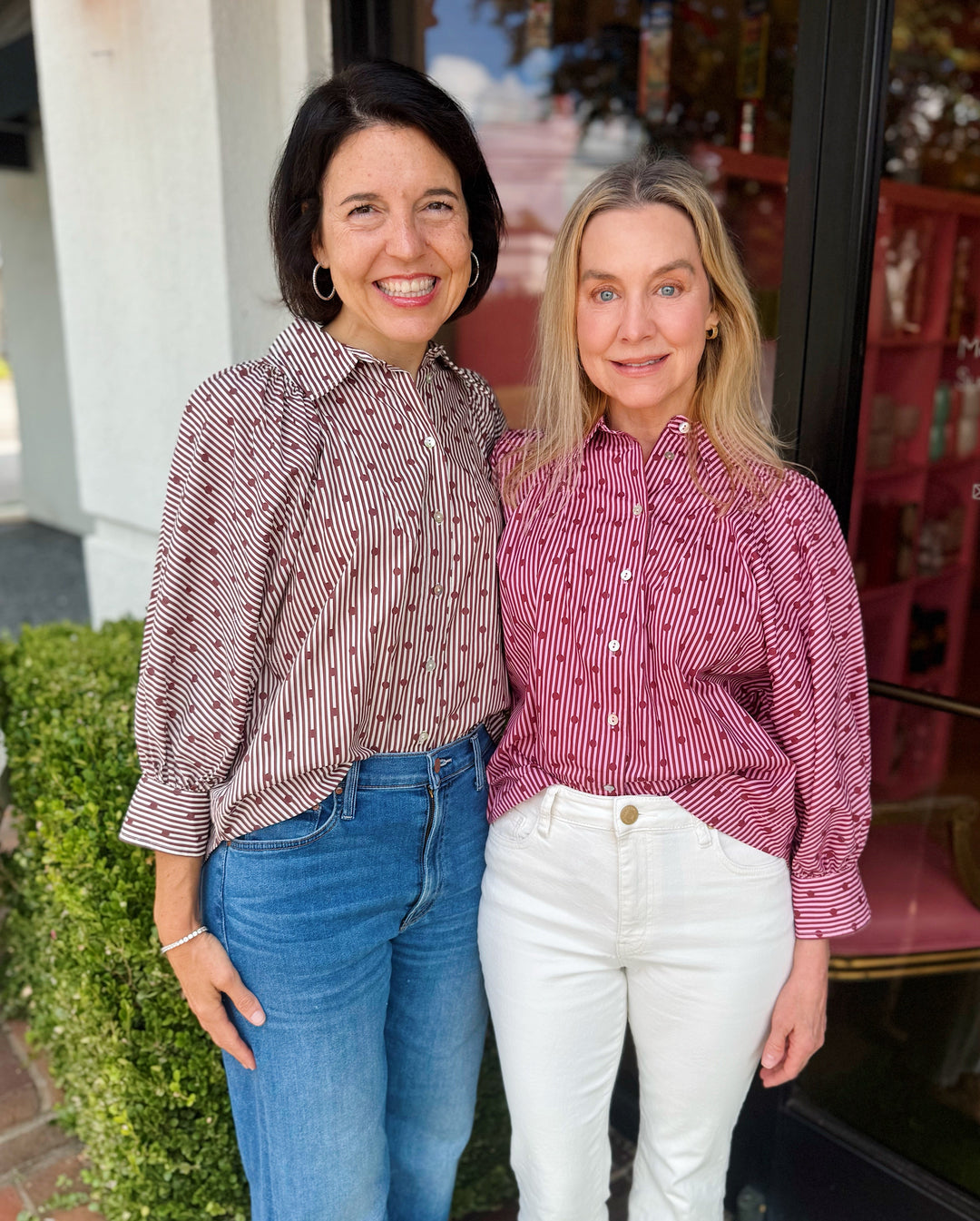 Two women standing side-by-side in front of a storefront window, wearing matching pink and white vertically striped button-down shirts.