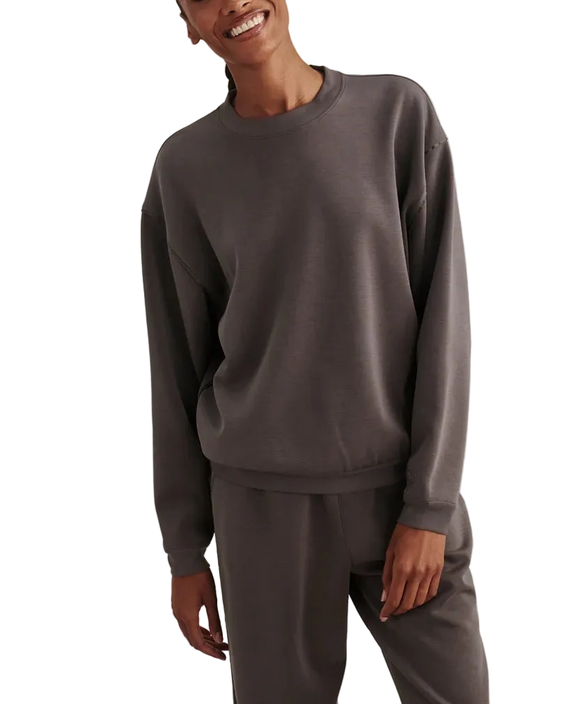A smiling woman wearing a dark gray crewneck sweatshirt and matching sweatpants, standing against a neutral textured beige background.