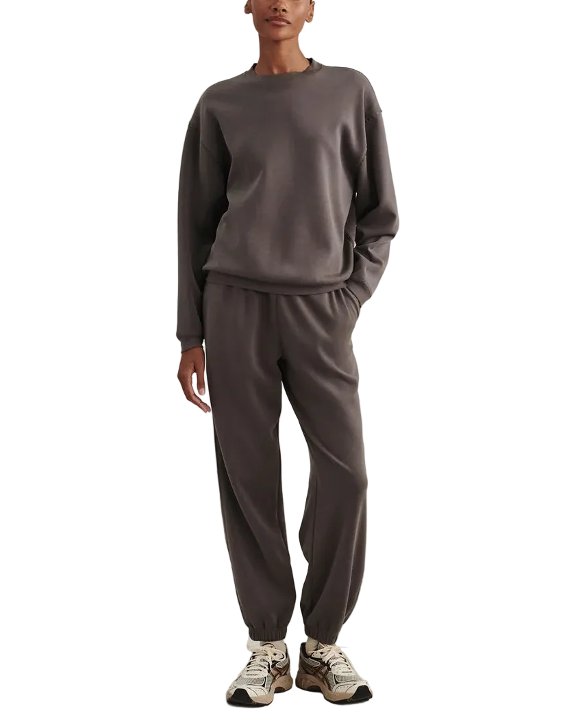 A model stands against a textured light beige studio backdrop wearing a matching dark gray crewneck sweatshirt and jogger pants set, paired with white and gray sneakers.