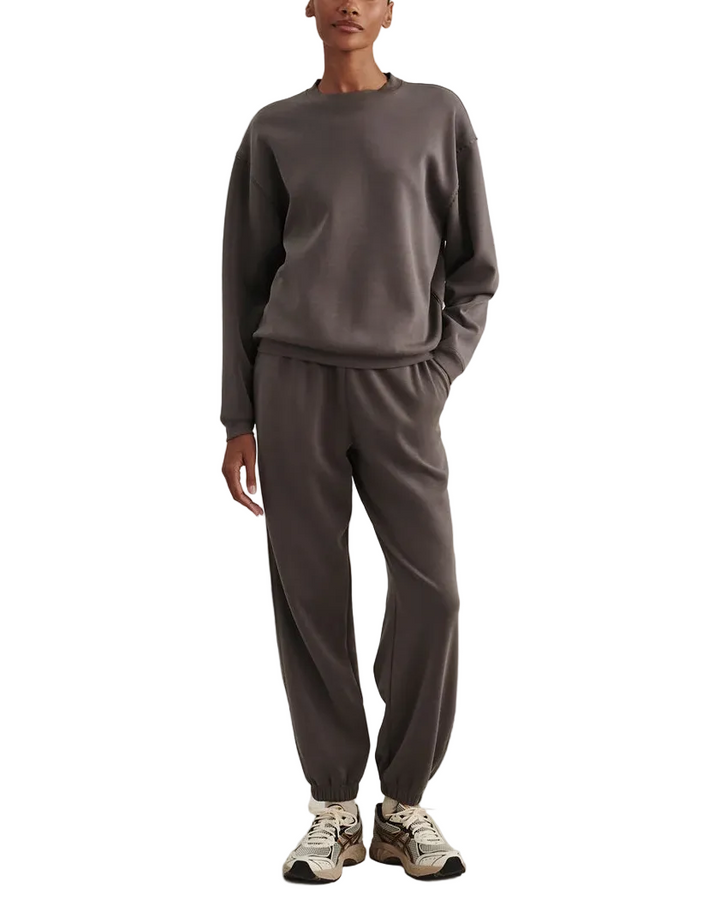 A model stands against a textured light beige studio backdrop wearing a matching dark gray crewneck sweatshirt and jogger pants set, paired with white and gray sneakers.