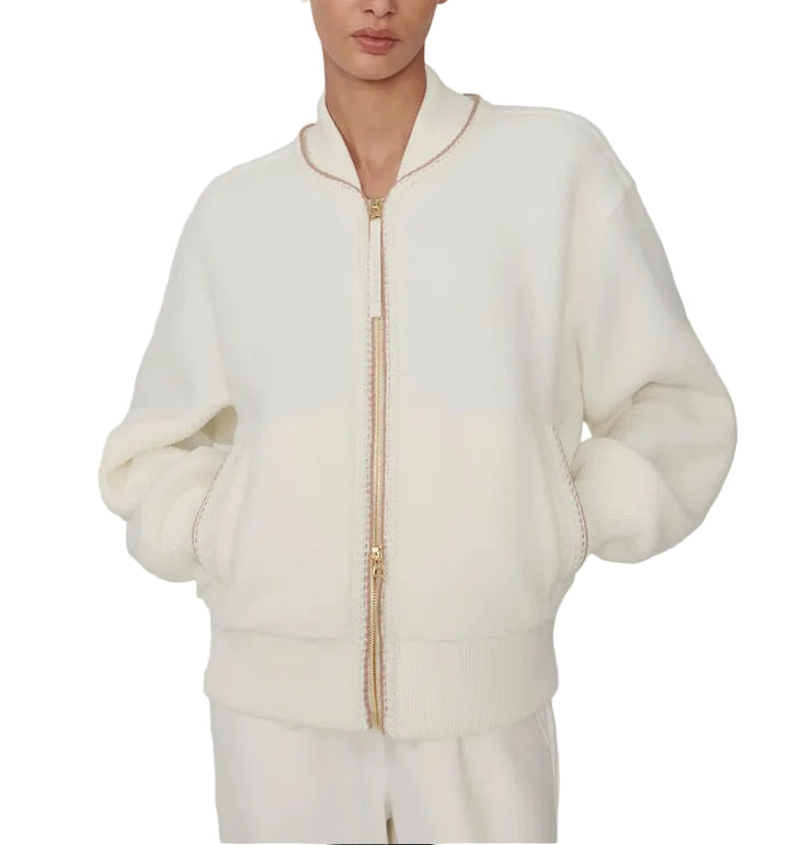 A model wearing a cream-colored fuzzy zip-up jacket with gold-tone zipper accents, paired with matching cream pants, against a plain light gray studio background.
