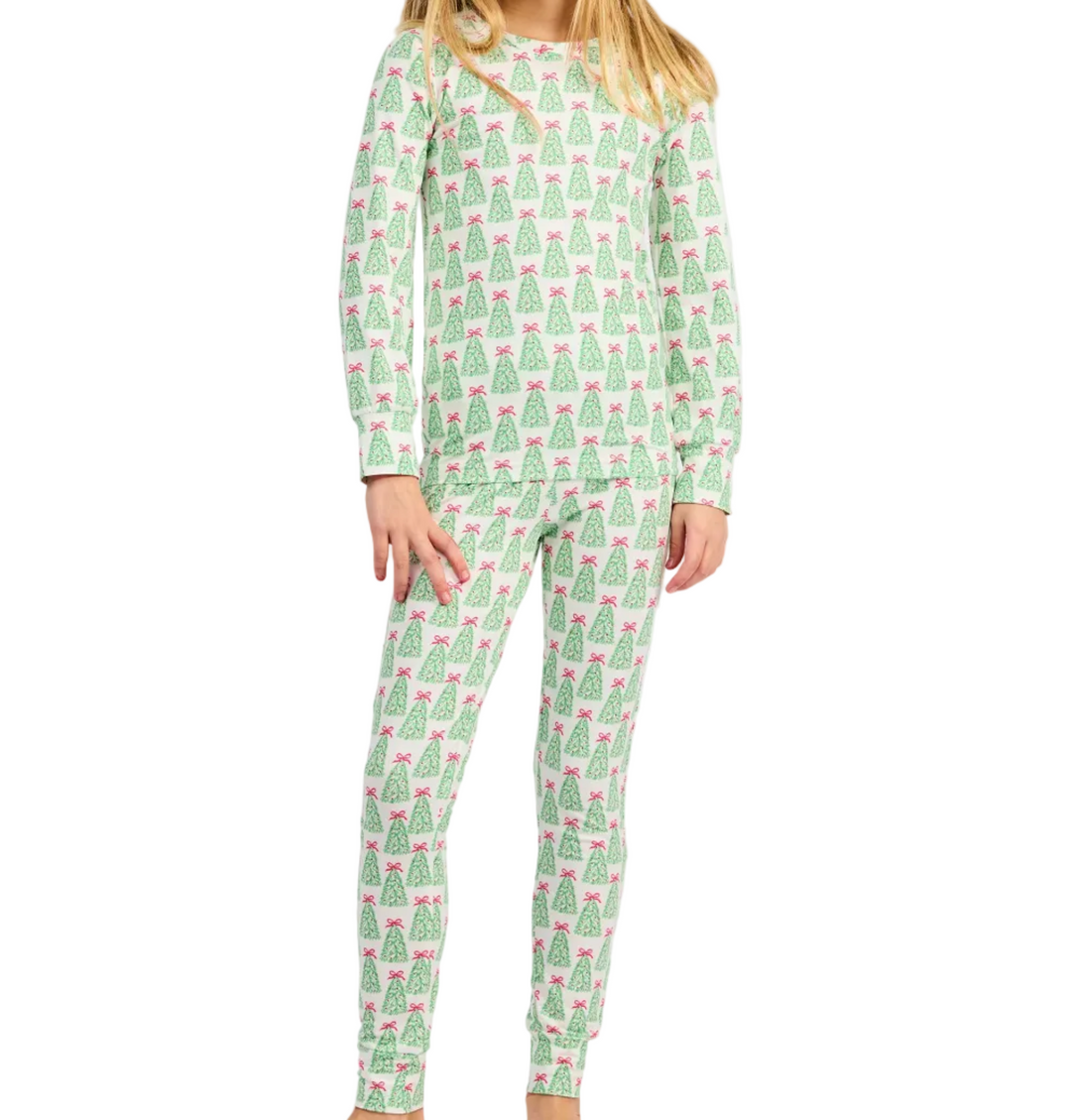 A young girl with blonde hair wearing a light green and white long-sleeve pajama set with a repeating Christmas tree pattern, standing against a plain white background.