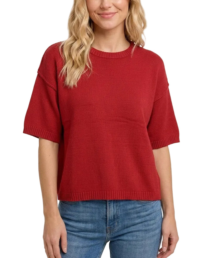 A woman with blonde wavy hair wearing a red short-sleeve knit top and blue jeans, standing against a plain light beige background.
