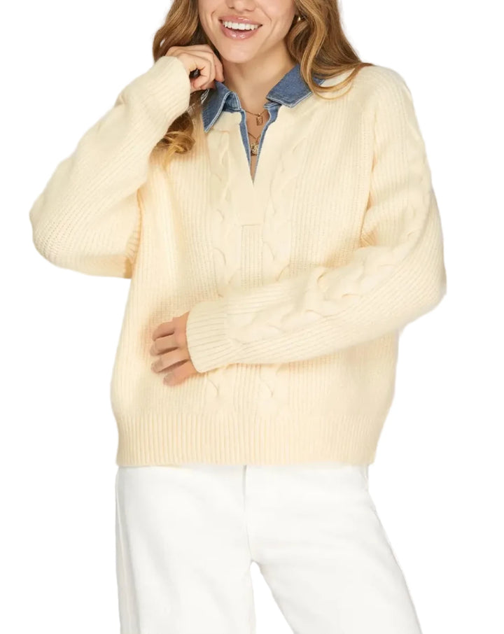 A woman wearing a cream cable-knit sweater over a blue denim collared shirt, paired with white pants, photographed against a light gray studio background.