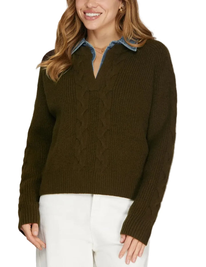 A woman wearing a dark olive green cable-knit sweater with a blue collar detail, paired with white pants, against a solid light gray background.
