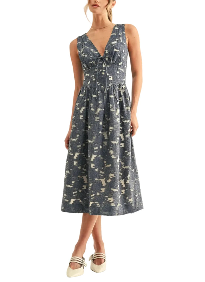 Cut from an intricate jacquard fabric in a denim blue jacquard colorway, it features a detailed floral pattern woven over a lighter, semi-sheer backing to create beautiful visual depth. The sleeveless bodice is designed with a deep V-neckline, gathered supportive cups, and a delicate tie detail at the center of the bust. Below the bust, a structured, corset-style waistband cinches the frame before flaring out into a voluminous, subtly pleated A-line midi dress