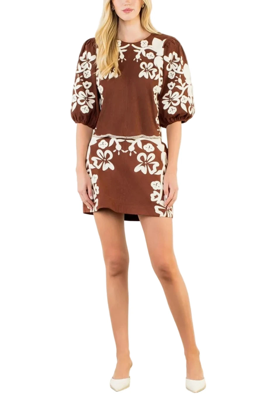A woman modeling a brown and white floral print mini dress with puff sleeves, standing against a plain white studio wall.