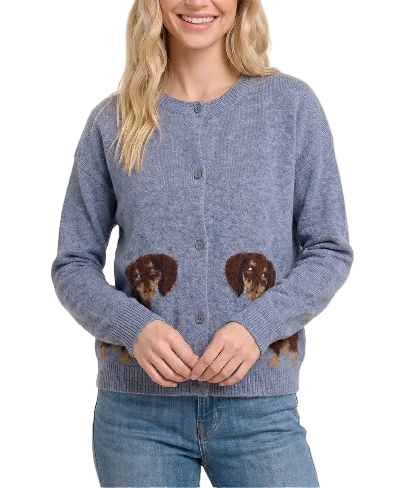 A smiling blonde woman wearing a heathered dusty blue button-up cardigan with embroidered dachshund dogs on the front, paired with blue jeans, against a solid light beige background.