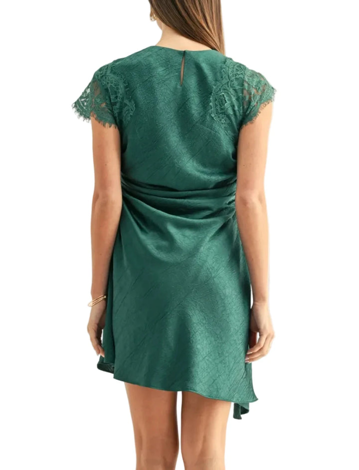 A woman viewed from the back wearing a short green satin dress with lace cap sleeves and a gathered waist detail, against a plain light background.