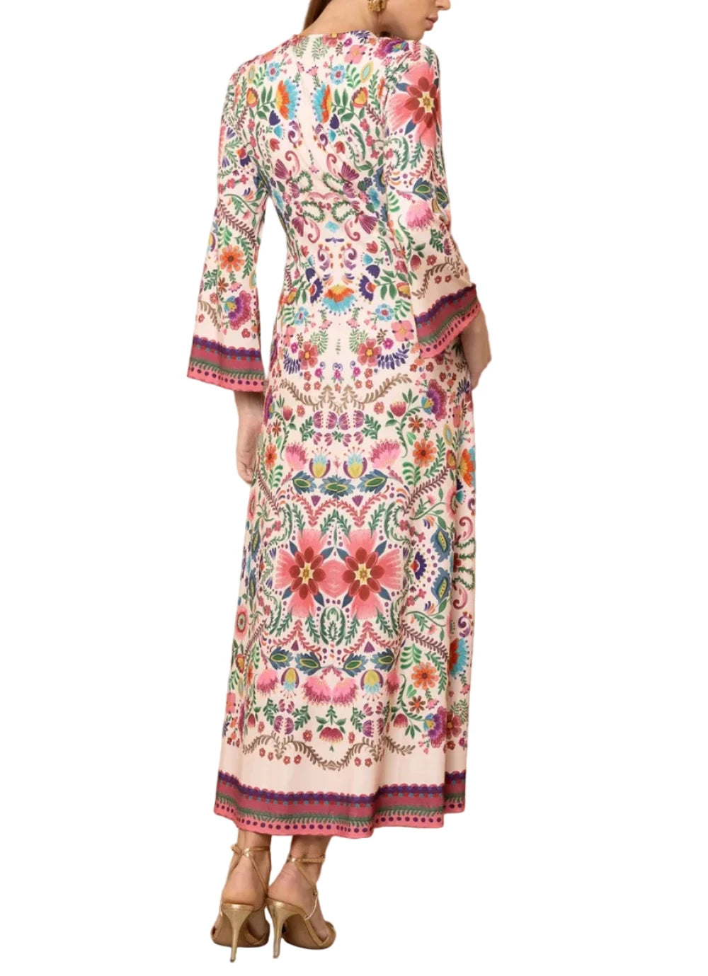 A woman modeling a long-sleeved maxi dress from the back, featuring a vibrant multicolor floral and geometric pattern on a cream base, set against a textured beige wall.