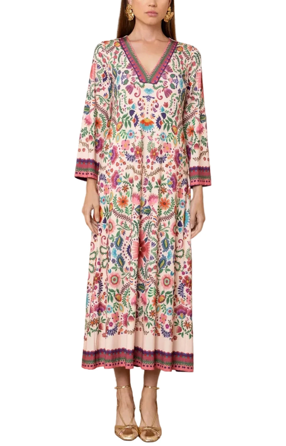 A woman models a long-sleeved, V-neck maxi dress with a vibrant multicolor floral print and embroidered borders, standing against a light beige textured wall.