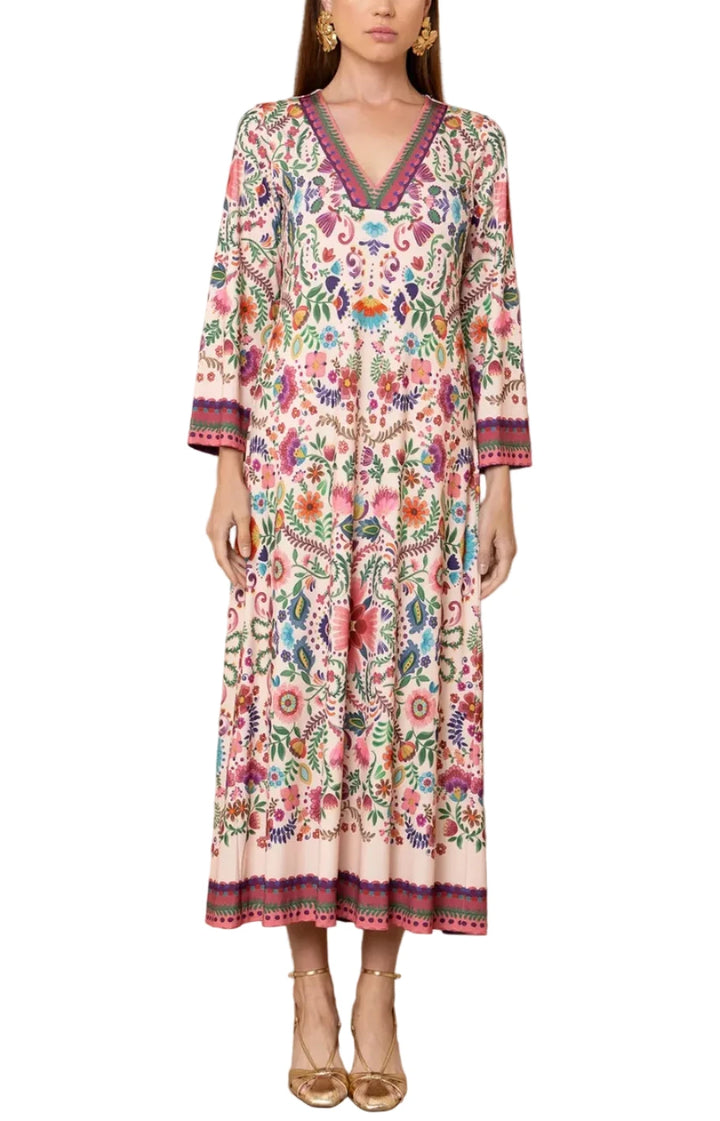 A woman models a long-sleeved, V-neck maxi dress with a vibrant multicolor floral print and embroidered borders, standing against a light beige textured wall.