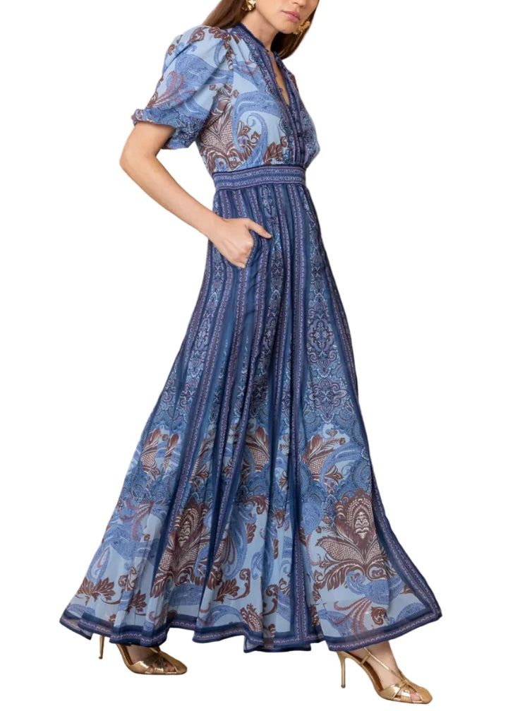 A woman models a long blue patterned maxi dress with puff sleeves, a V-neckline, and a pleated skirt, standing against a textured beige wall.