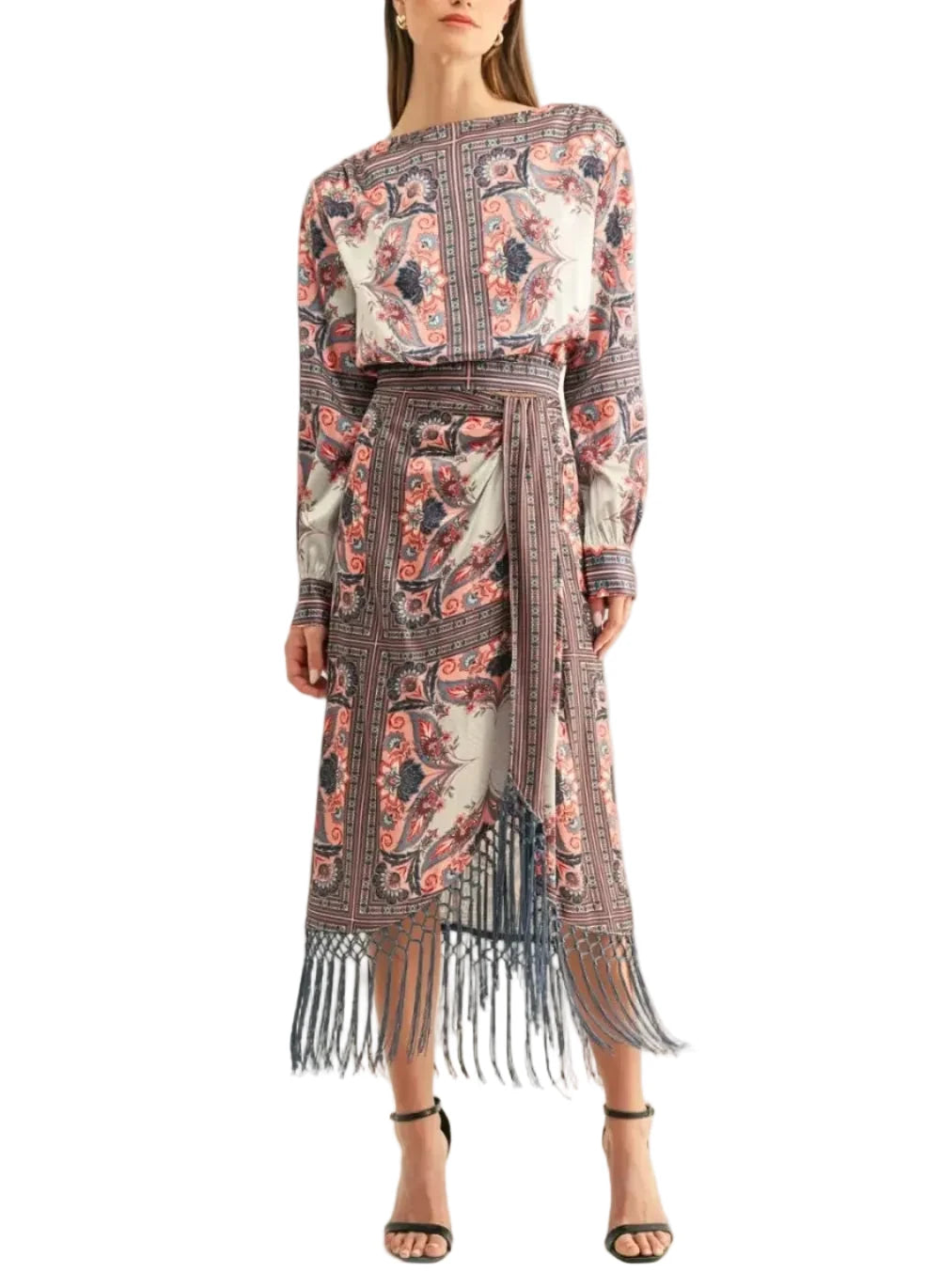 A woman modeling a long-sleeved midi dress with an intricate multicolor paisley and floral print, featuring a matching self-tie belt and a long fringe hem, set against a plain off-white studio background.
