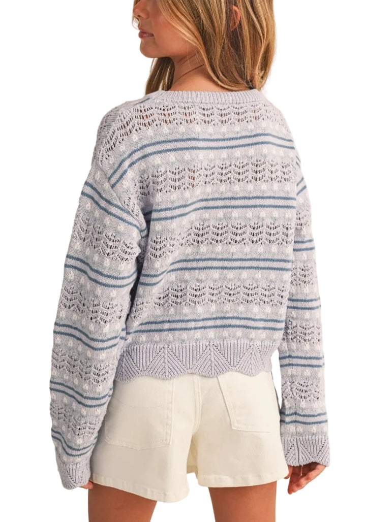 A girl shown from the back wearing a light blue and white patterned knit sweater and white shorts against a solid beige background.