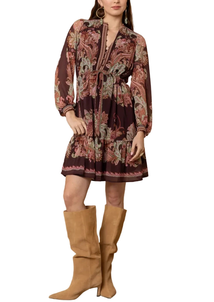 A woman modeling a long-sleeve, knee-length dress with a paisley pattern in deep burgundy, brown, and beige, paired with tan knee-high boots, standing against a textured beige wall.