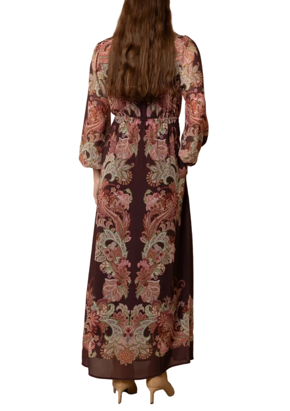 A woman viewed from behind wearing a long-sleeved maxi dress with a detailed paisley pattern in dusty pink, rust, and beige over a dark plum base, standing against a textured beige wall.