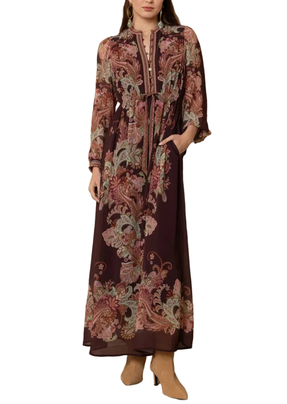 A woman modeling a long-sleeved maxi dress with a dark brown base and an intricate paisley floral print in dusty pink, beige, and muted green. The dress features a high collar, buttoned front, and a self-tie belt at the waist, paired with tan ankle boots against a textured beige wood-paneled background.