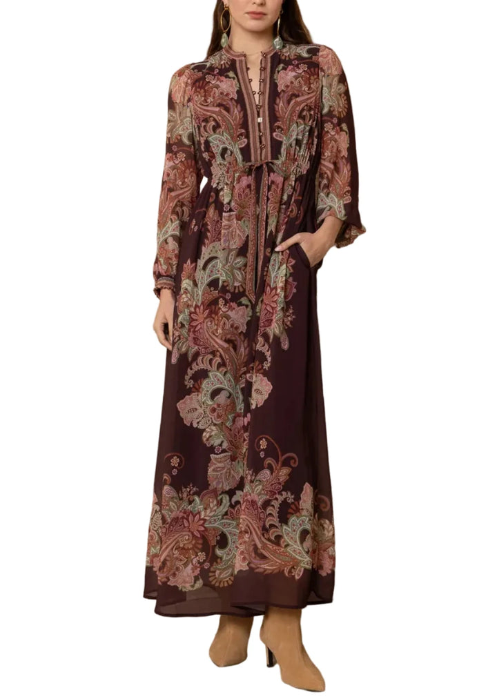 A woman modeling a long-sleeved maxi dress with a dark brown base and an intricate paisley floral print in dusty pink, beige, and muted green. The dress features a high collar, buttoned front, and a self-tie belt at the waist, paired with tan ankle boots against a textured beige wood-paneled background.