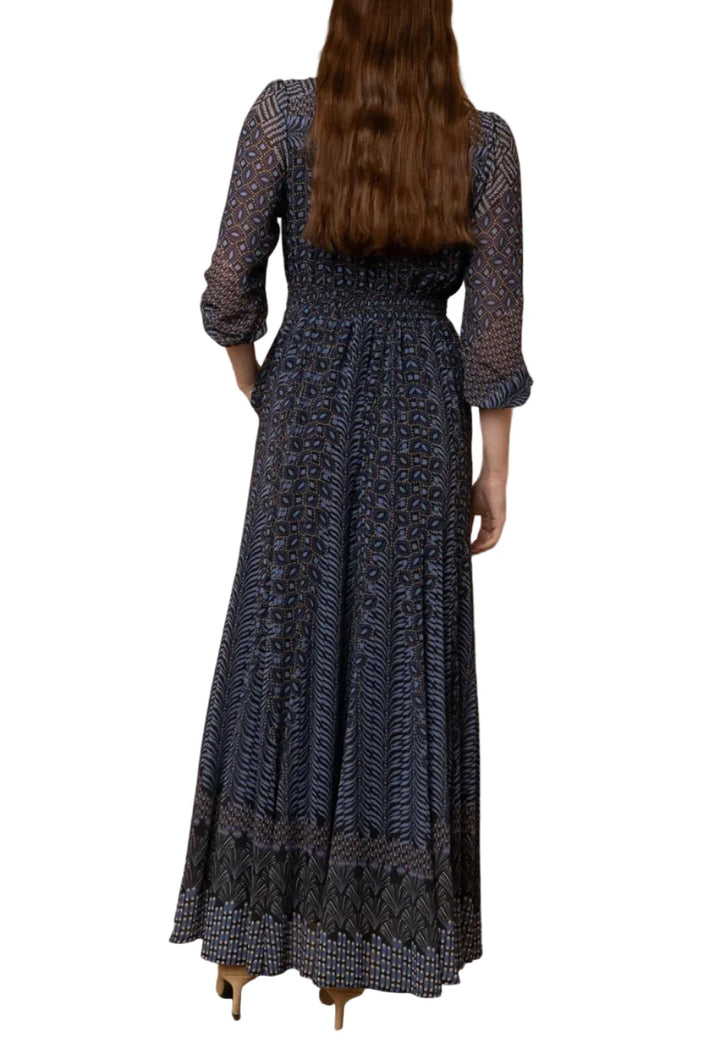 A woman viewed from behind wearing a long navy maxi dress with a detailed geometric pattern, standing against a light wood-paneled wall.