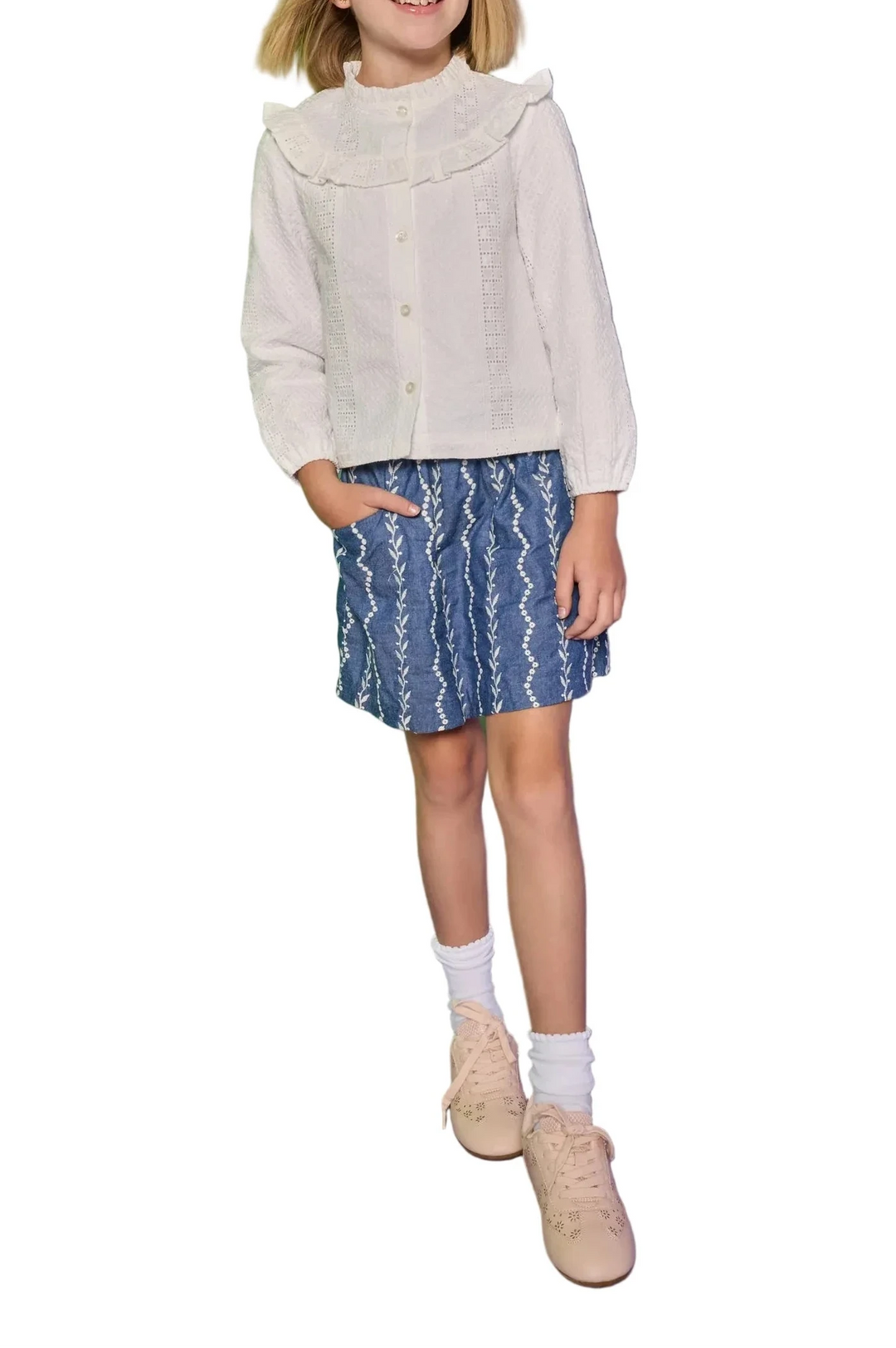 Young girl in a light gray jacket and blue patterned skirt standing against a green and purple background