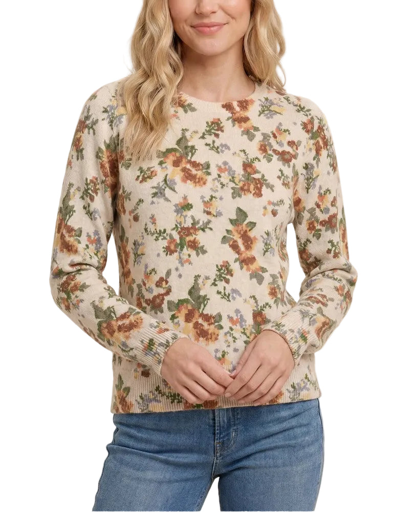 A woman wearing a beige long-sleeve crewneck sweatshirt with an all-over floral print in orange, green, and brown, paired with blue jeans, against a solid cream background.