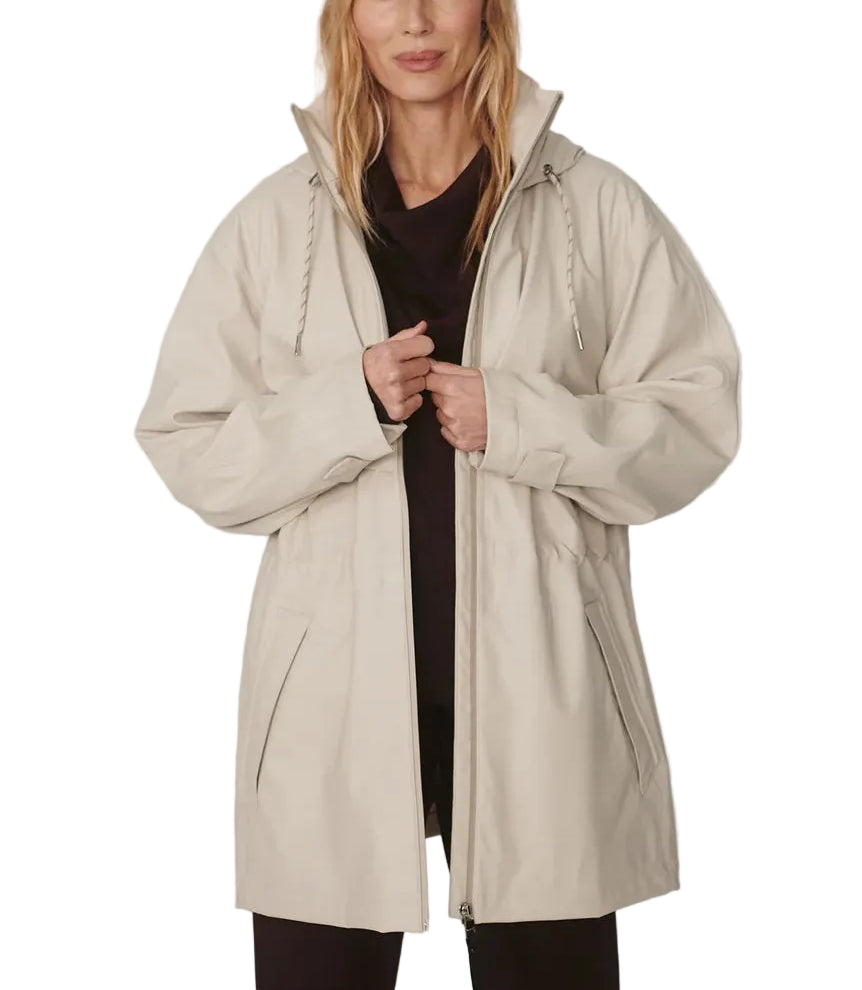A woman modeling a light beige hooded raincoat over a black top and black pants, wearing a black baseball cap with a white logo, against a plain light gray studio background.