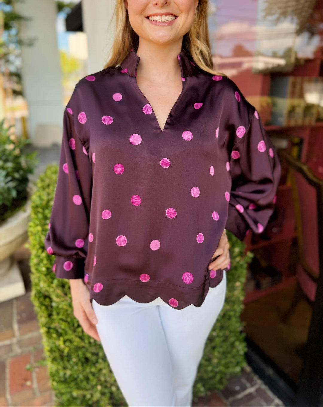 A woman wearing a dark purple satin blouse with pink polka dots and a scalloped hem, paired with white pants, standing outdoors in front of a building.