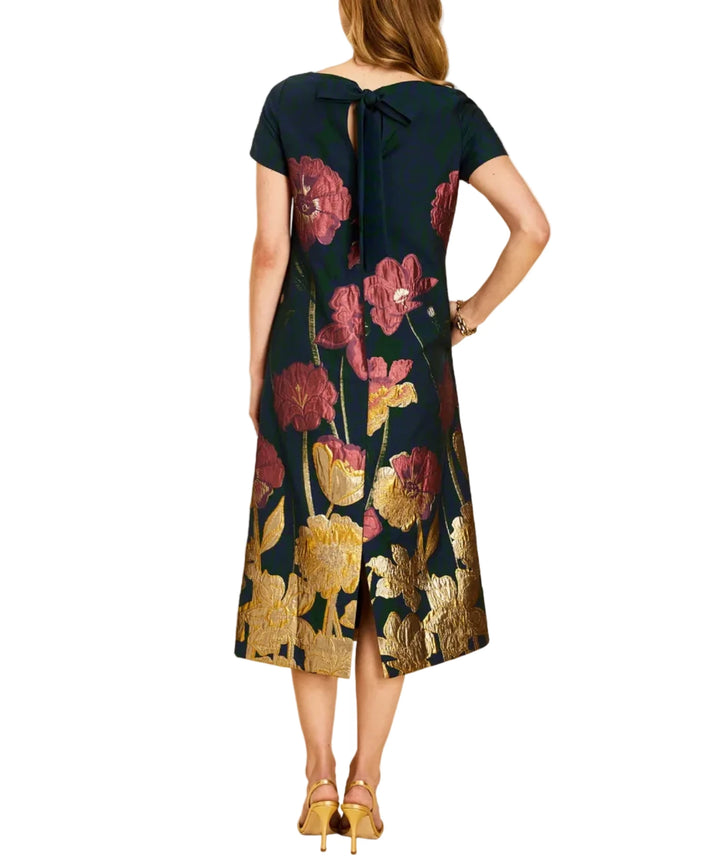 A woman shown from the back wearing a short-sleeve midi dress with a bow at the neckline and a dark floral print with pink, red, and gold flowers, paired with gold heels, against a solid beige background.