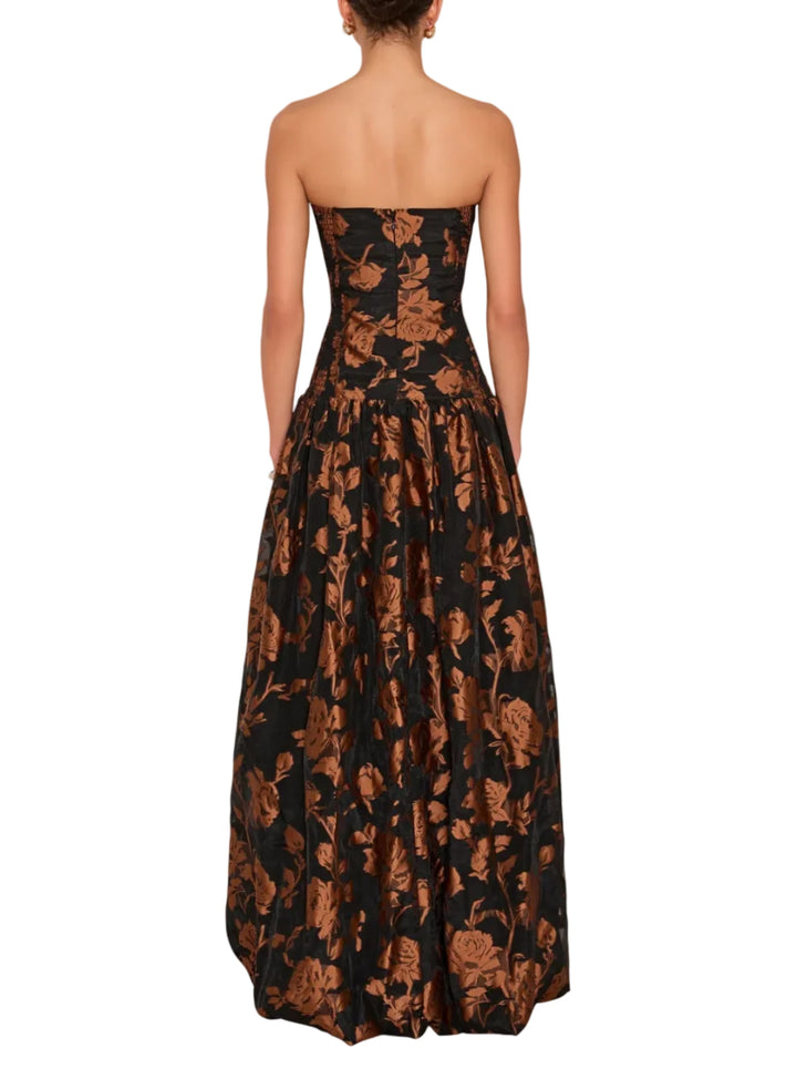 A strapless floor-length gown shown from the back on a model, featuring a black base with a metallic bronze floral pattern.