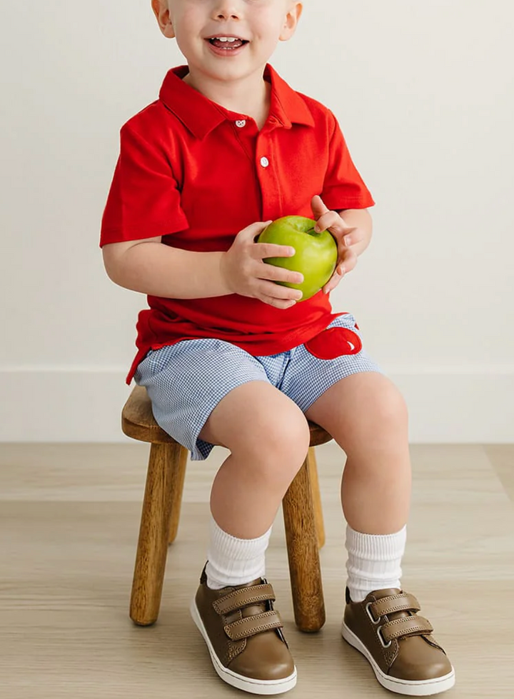 A smiling toddler sits on a small wooden stool wearing a bright red short-sleeve polo shirt, light blue and white striped shorts, white socks, and brown velcro shoes, while holding a green apple.