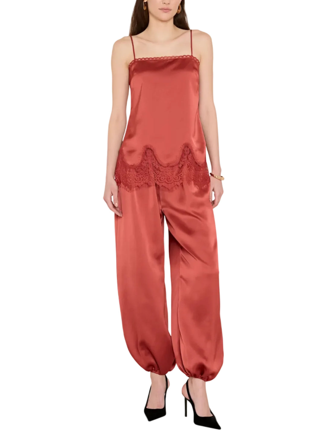 A woman modeling a rust-colored satin jumpsuit with thin straps, a lace-trimmed hem, and wide baggy legs, paired with black pointed-toe heels against a light gray studio background.