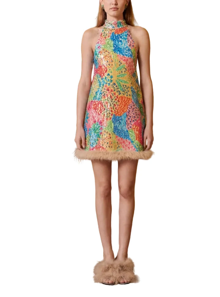 A model stands in a studio setting wearing a sleeveless, high-neck mini dress covered in a vibrant multicolor abstract pattern with a fuzzy beige trim at the hem. The background features a warm mustard yellow wall and a wooden panel on the left, with a brown leather chair partially visible.