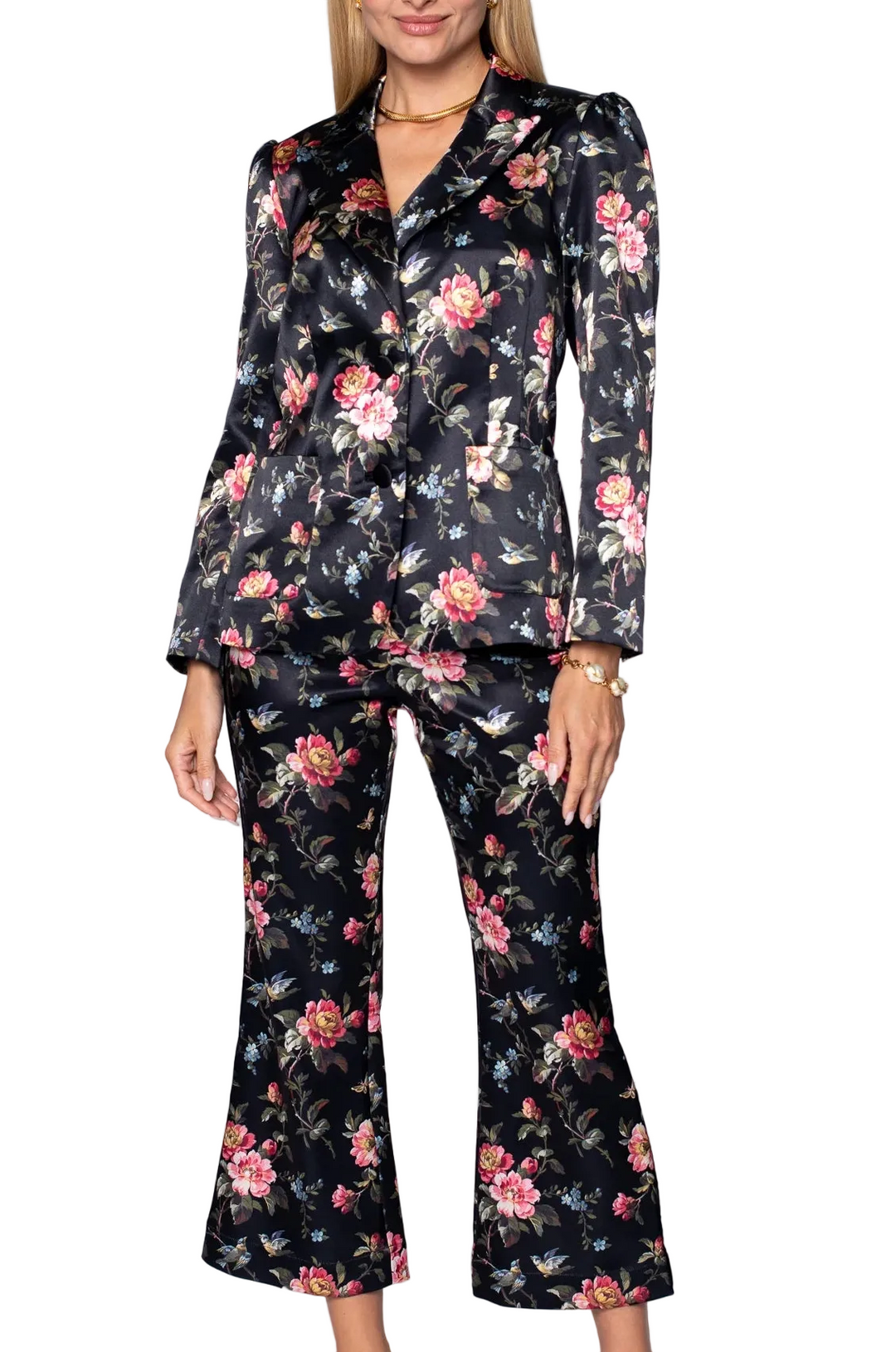 A woman modeling a black satin-like two-piece suit with a pink and red floral print, featuring a blazer and matching cropped pants, against a light gray studio background.