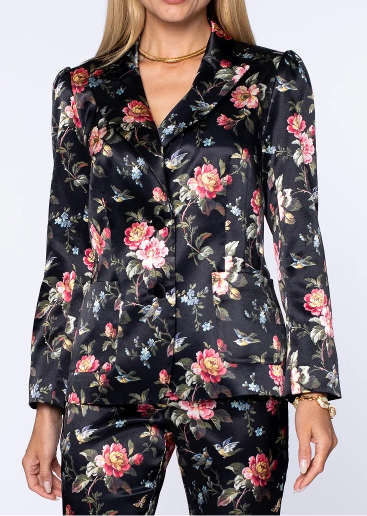 A woman modeling a matching black floral satin blazer and trousers set with pink, red, and blue flowers, set against a plain light gray studio background.