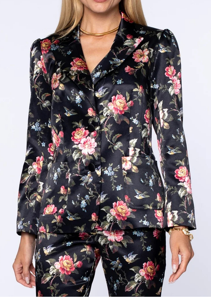 A woman modeling a matching black floral satin blazer and trousers set with pink, red, and blue flowers, set against a plain light gray studio background.