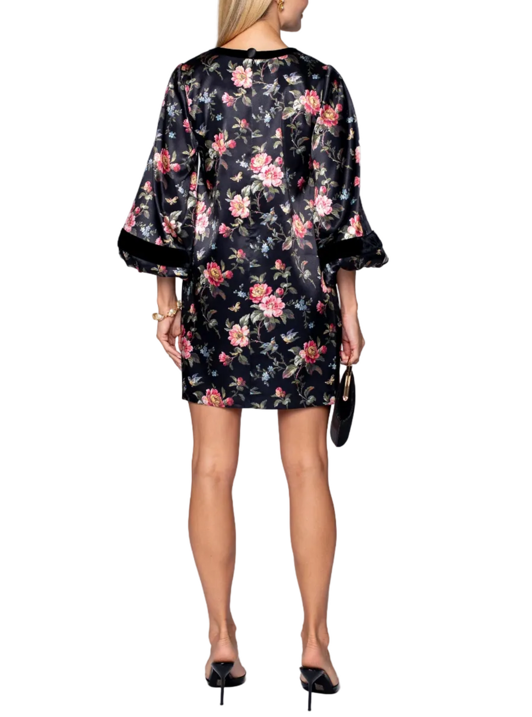 A woman viewed from the back wearing a short black satin-like dress with a pink and green floral print, wide three-quarter sleeves with black trim, and holding a small black handbag against a light gray studio background.