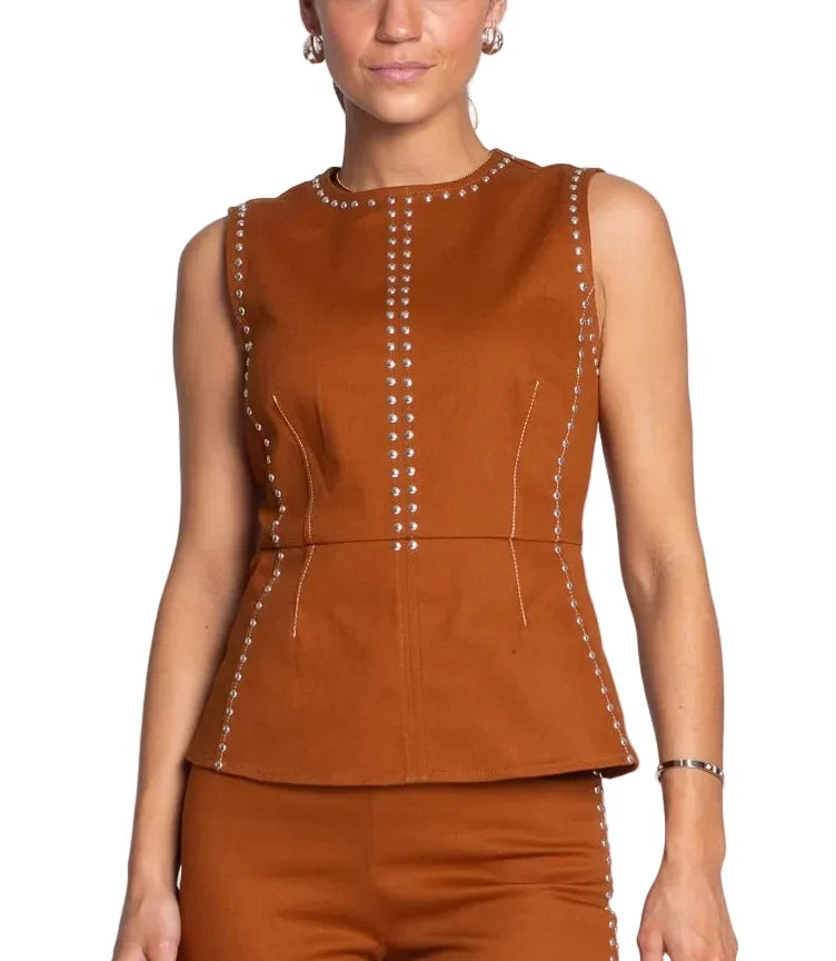A woman modeling a sleeveless rust-brown peplum top with silver stud detailing along the neckline, seams, and front, paired with matching rust-brown pants against a light gray studio background.