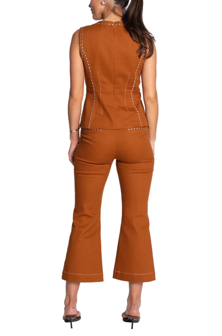 A woman viewed from the back wearing a rust-orange sleeveless top and matching flared pants with contrast stitching and studded details, photographed against a light gray studio background.