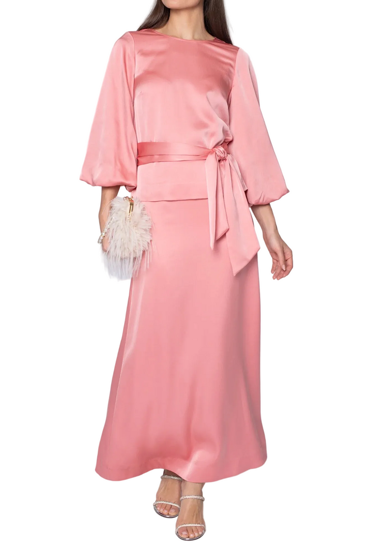 A woman modeling a satin-like pink maxi dress with three-quarter sleeves, a matching waist tie, and a flared skirt, standing against a light gray studio background.