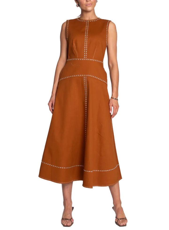 A woman modeling a sleeveless rust-brown midi dress with white embroidered trim details, standing against a light gray studio background.