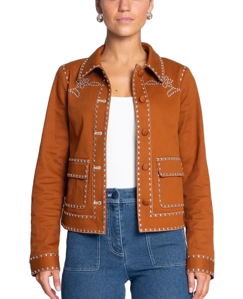A woman modeling a cropped brown suede jacket with white embroidered details and silver-toned studs, worn over a white top and blue jeans against a light gray background.