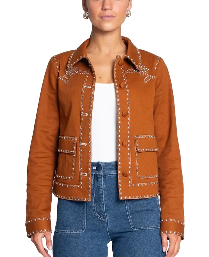 A woman modeling a cropped brown suede jacket with white embroidered details and silver-toned studs, worn over a white top and blue jeans against a light gray background.