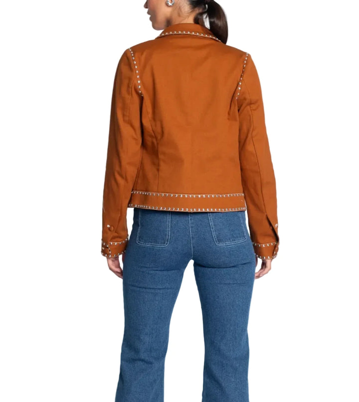 A woman shown from the back wearing a rust-orange cropped jacket with metallic stud detailing and blue flare jeans, photographed against a light gray studio background.